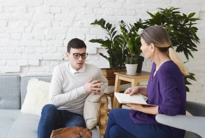candid-shot-young-man-glasses-talking-about-his-problems-during-psychological-therapy-session-sitting-coach-while-mature-female-psychologist-with-copybook-listening-him-making-notes_344912-1939